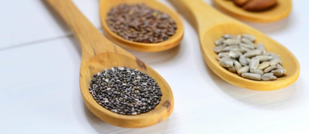 A variety of seeds in wooden spoons displayed on a white surface.